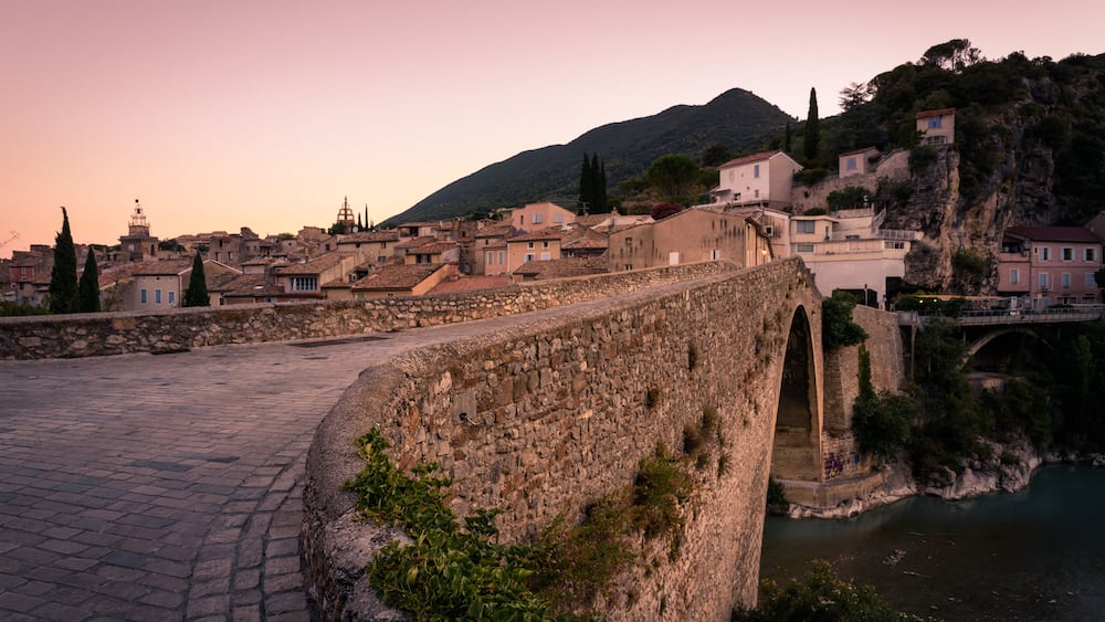Un pont de pierre, une rivière et une ville avec des bâtiments, le tout surmonté d’une colline en arrière-plan.