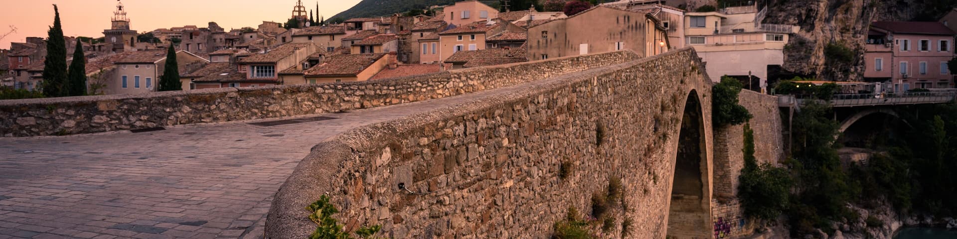 A stone bridge, a river, and a town with buildings and a hill in the background.