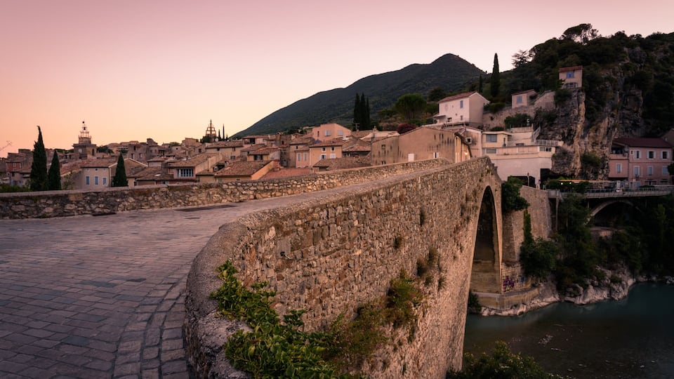 A stone bridge, a river, and a town with buildings and a hill in the background.