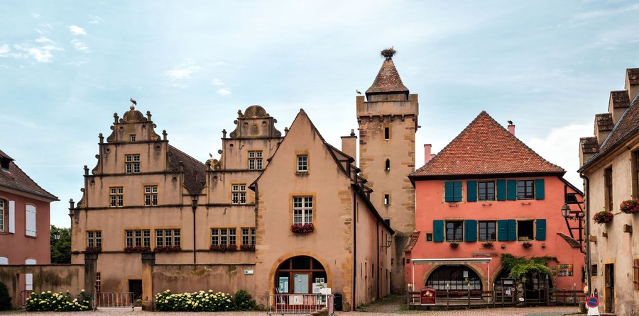 En historisk europeisk stad med en framträdande byggnad som liknar ett slott och ett färgglatt hus med rött tak.