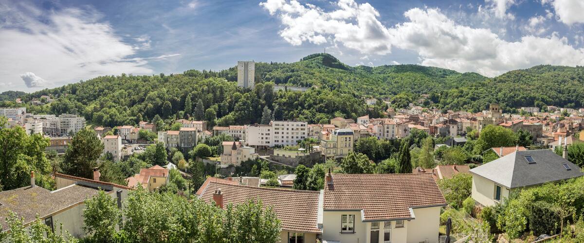 A town with red-tiled roofs nestled in a valley, surrounded by lush green hills and a prominent hill with a tower.