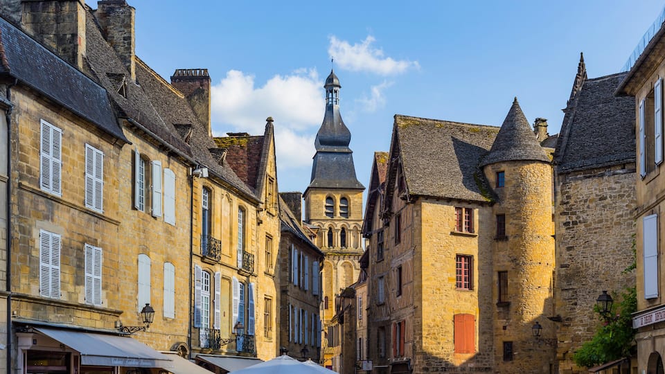 A narrow street lined with historic buildings, a prominent tower, and a market area with stalls.