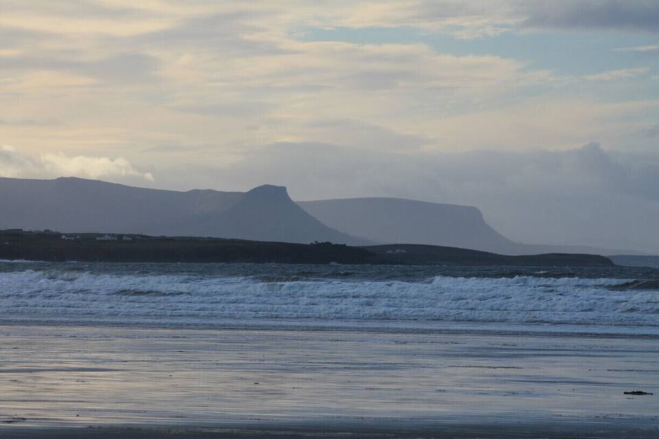 Una spiaggia con onde e montagne in lontananza, sotto un cielo nuvoloso.