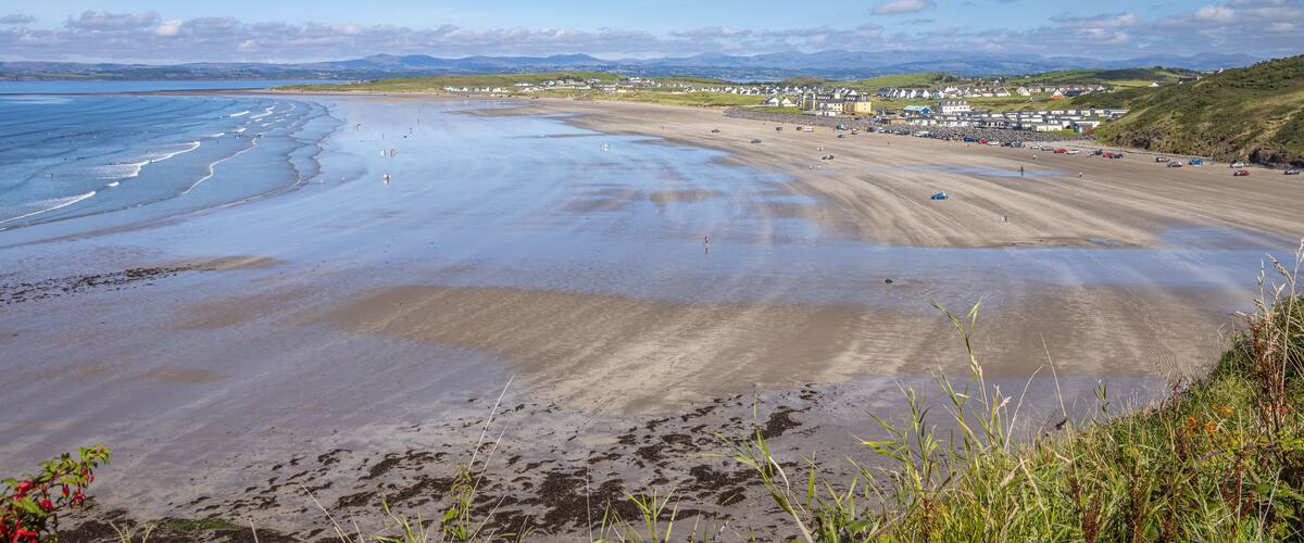 A wide sandy beach with gentle waves, a small boat, and a town in the background.