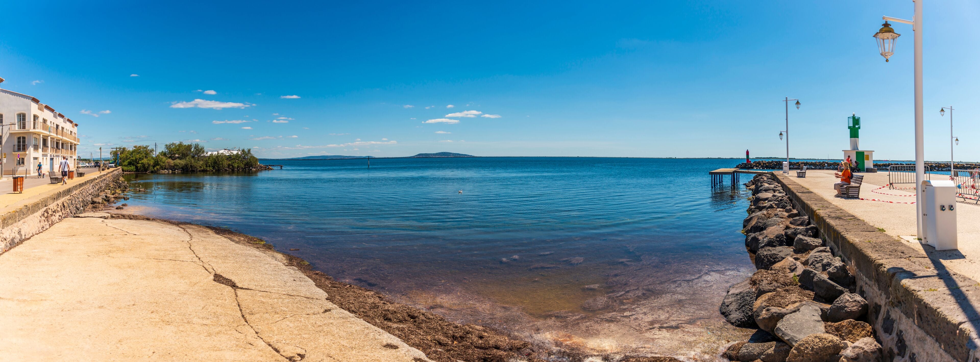 Paisaje costero con cielo azul despejado, agua tranquila, un rompeolas rocoso y una isla distante.