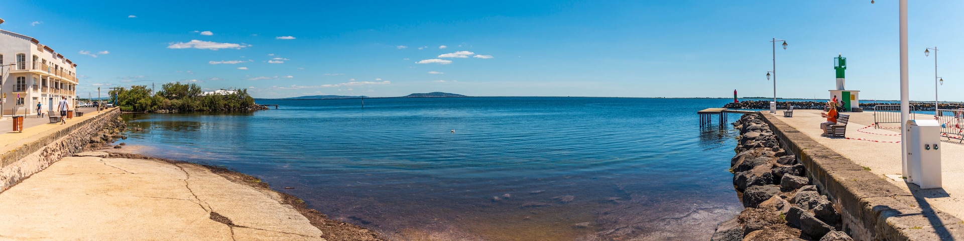 Paisaje costero con cielo azul despejado, agua tranquila, un rompeolas rocoso y una isla distante.