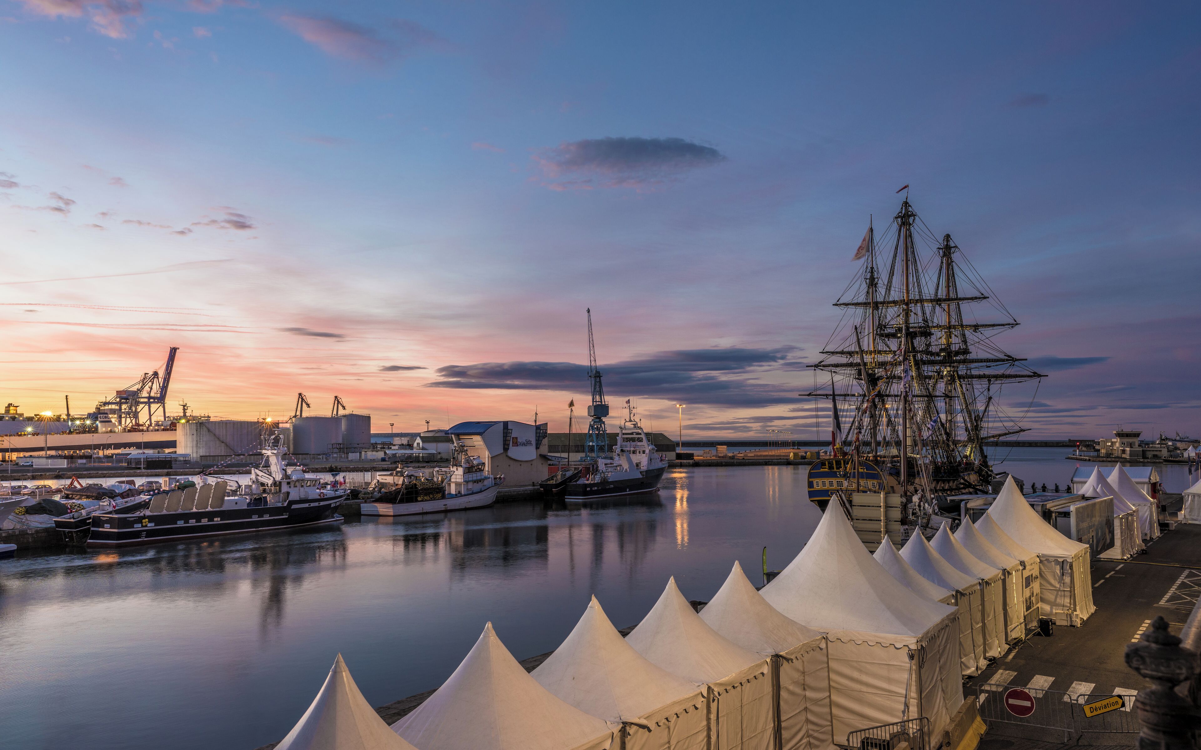 A harbor at sunset with a tall ship, boats, and tents.