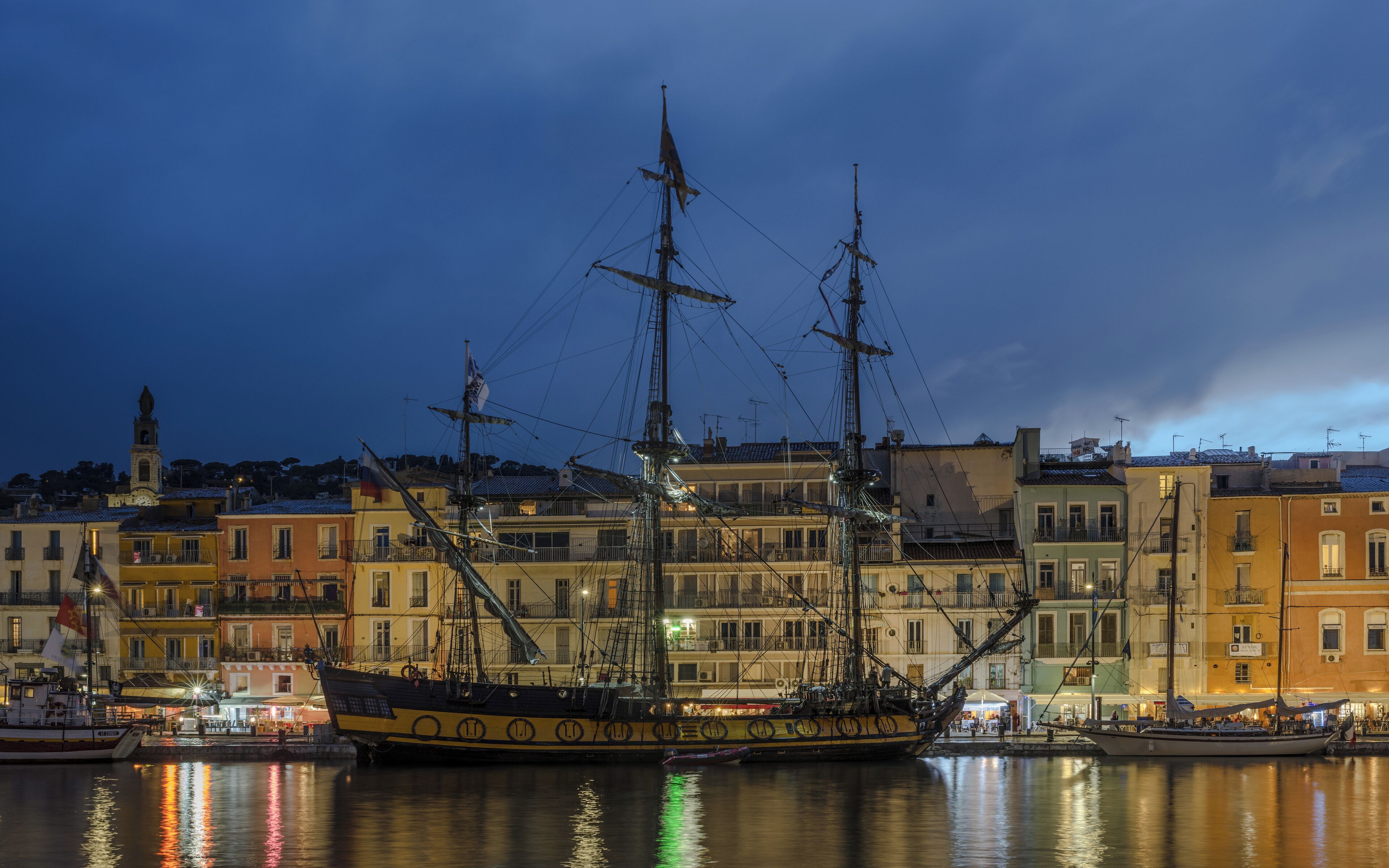 A large sailing ship docked in a harbor at night, with colorful buildings in the background.