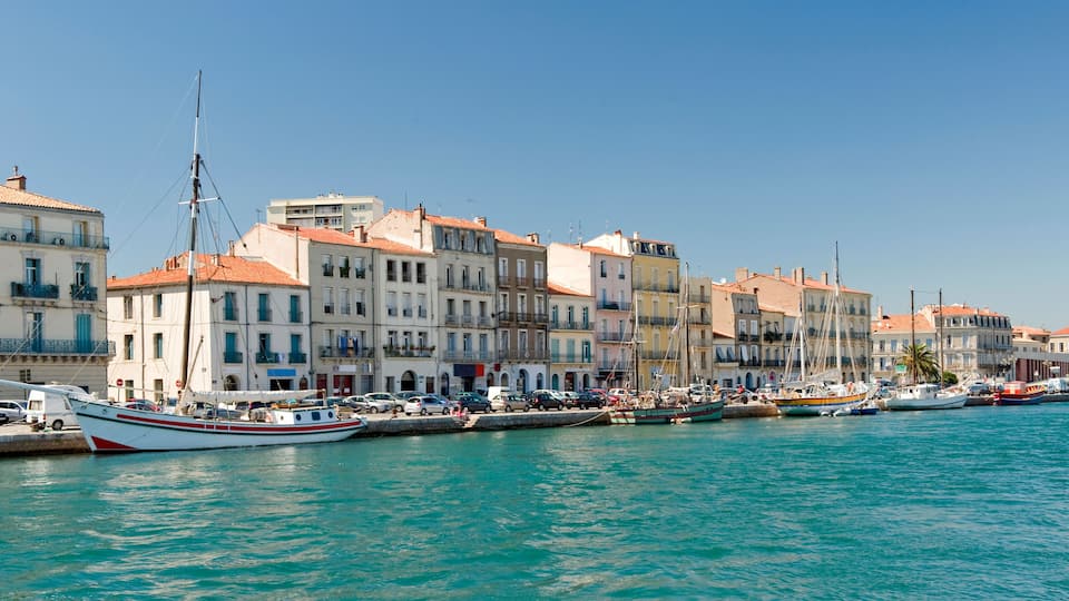 A waterfront with boats docked, buildings with red-tiled roofs, and a clear blue sky.