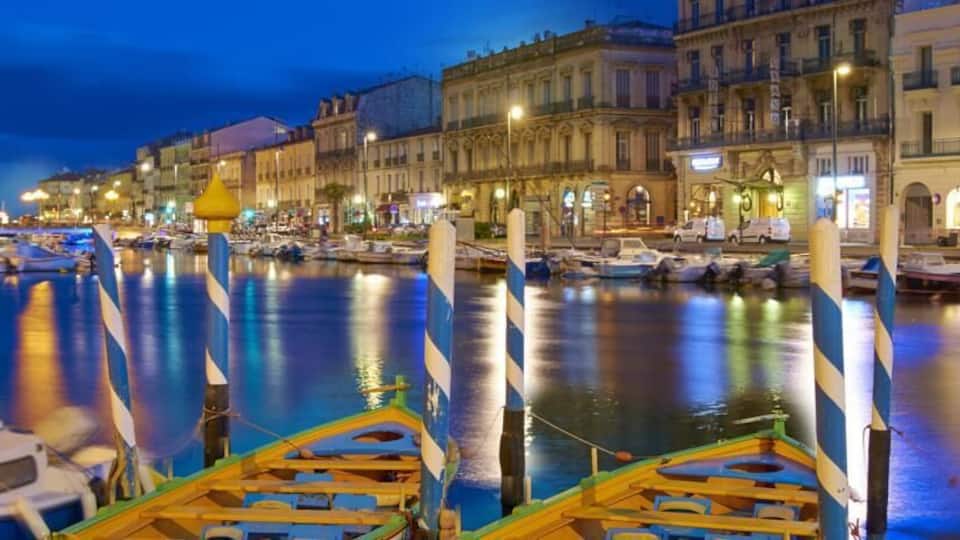 A marina at dusk with boats docked, striped bollards, and illuminated buildings along the waterfront.