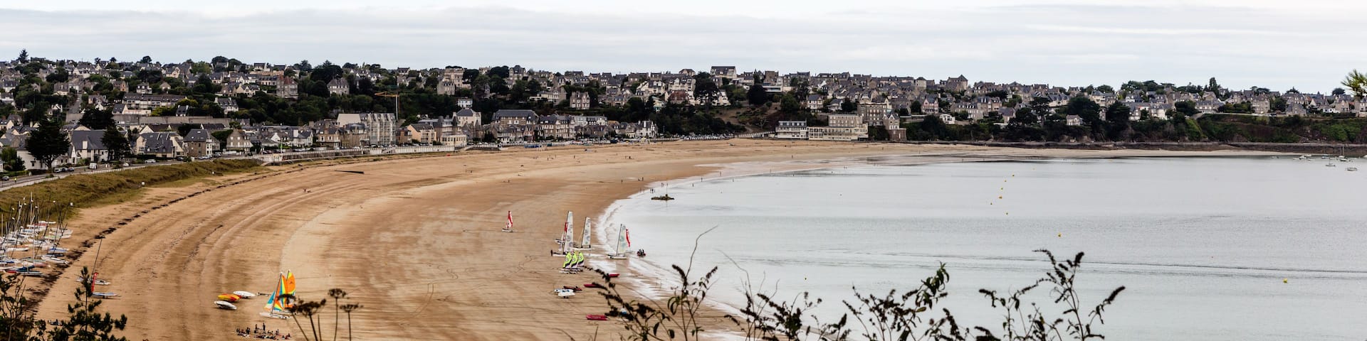 A coastal town with a sandy beach, sailboats, and a backdrop of residential buildings.