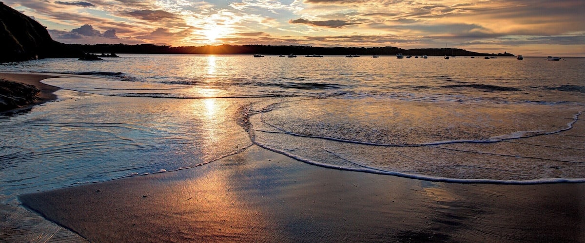 A beach at sunset with the sun reflecting off the water, waves, and a distant shoreline with boats.