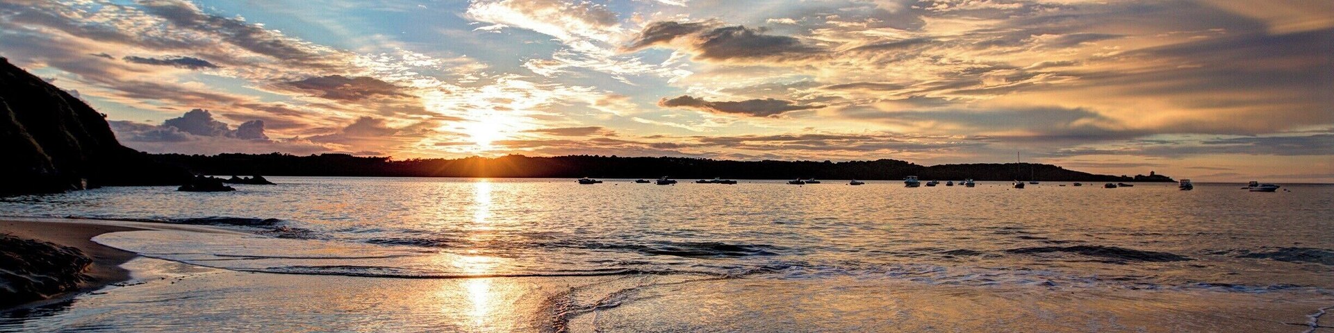 A beach at sunset with the sun reflecting off the water, waves, and a distant shoreline with boats.