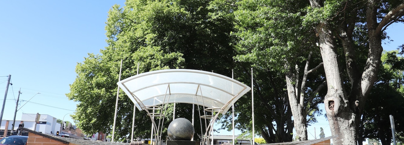 Placa acolhedora com toldo e uma escultura esférica no centro, cercada por calçada de tijolos e vegetação.