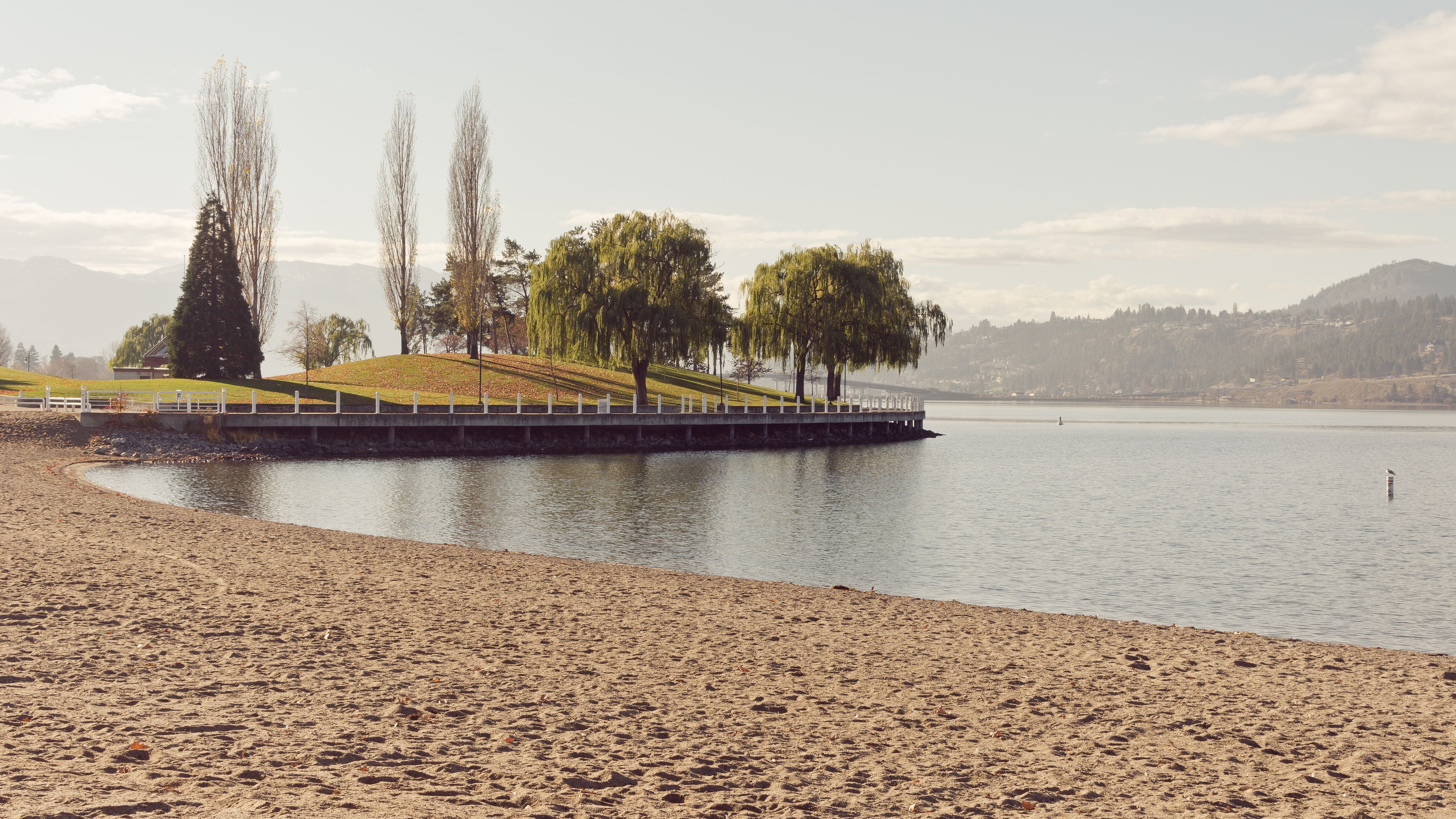 A sandy beach with a wooden pier, trees, and a calm lake.