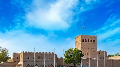 Una fortaleza histórica con una torre destacada, rodeada de exuberante vegetación y un cielo azul despejado.
