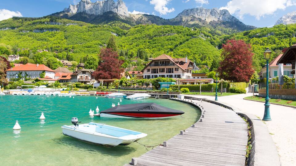 A marina with boats docked, a wooden pier, and houses in a mountainous area.