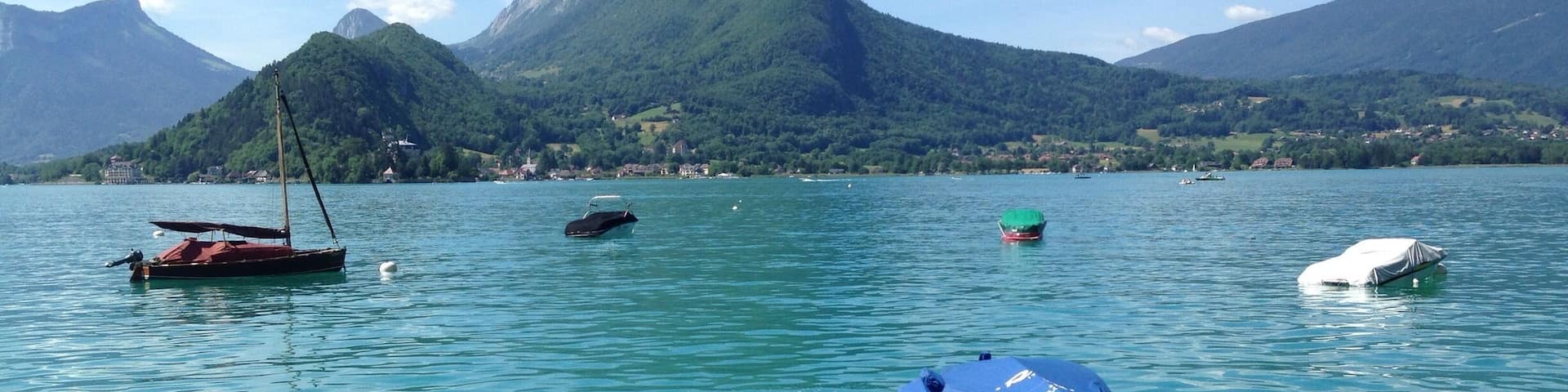 A calm lake with several boats, a sailboat, and a covered dinghy. In the background, there are mountains and a small village.
