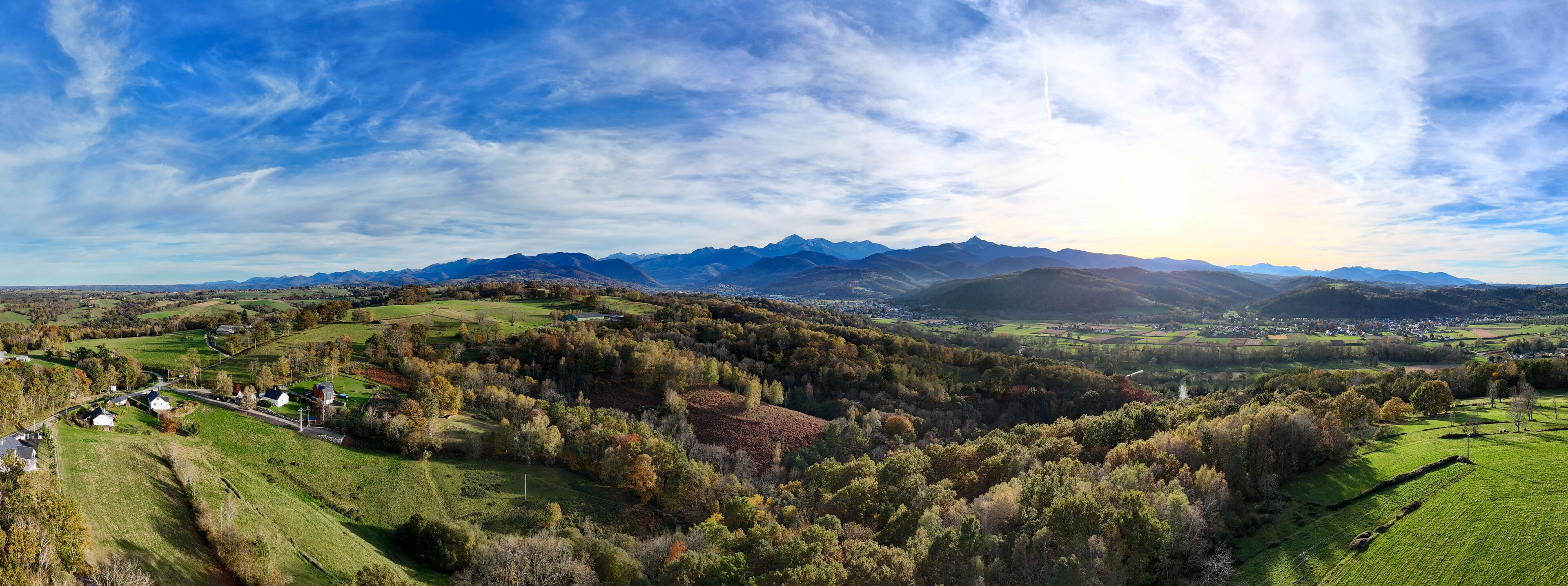 A panoramic view of a valley with rolling hills, a river, and distant mountains under a partly cloudy sky.