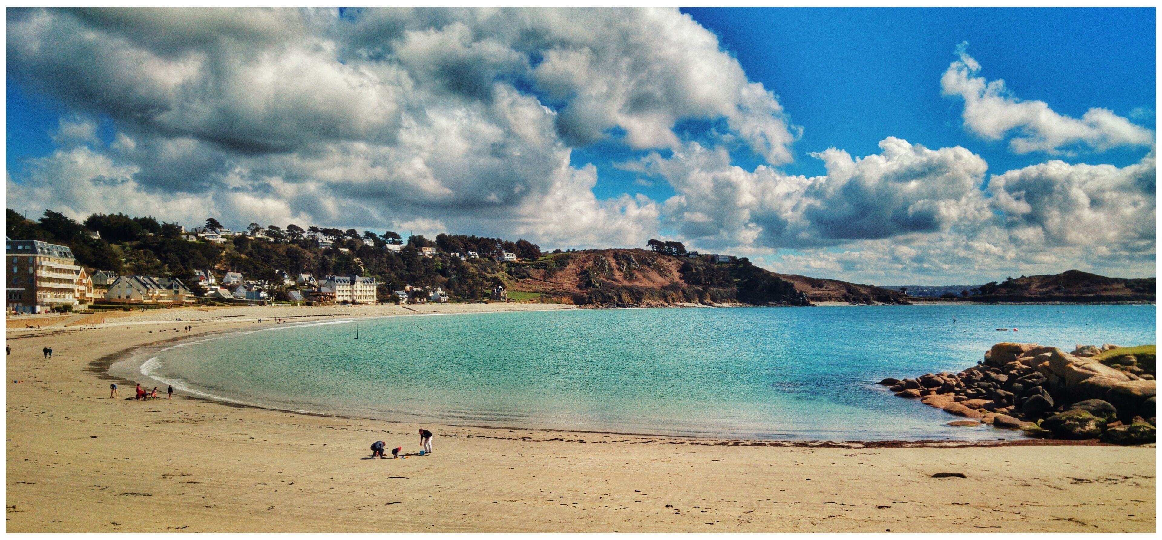 A beach with clear turquoise water, a sandy shore, and houses on a hillside in the background.