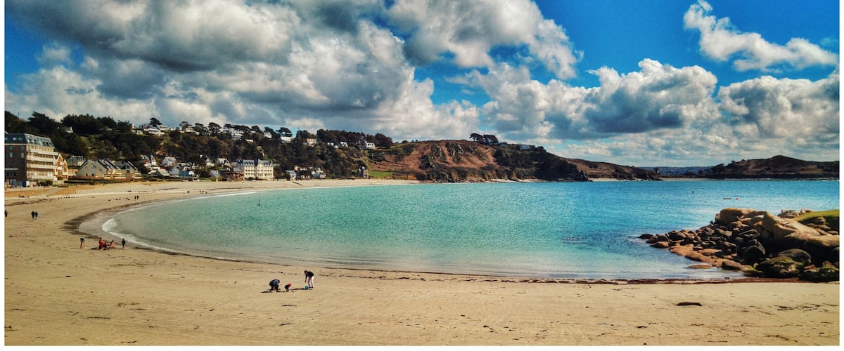 A beach with clear turquoise water, a sandy shore, and houses on a hillside in the background.