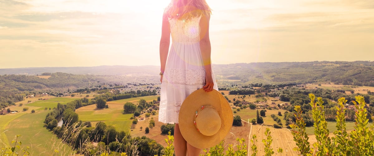A woman in a white dress and straw hat overlooking a rural landscape with fields and trees.
