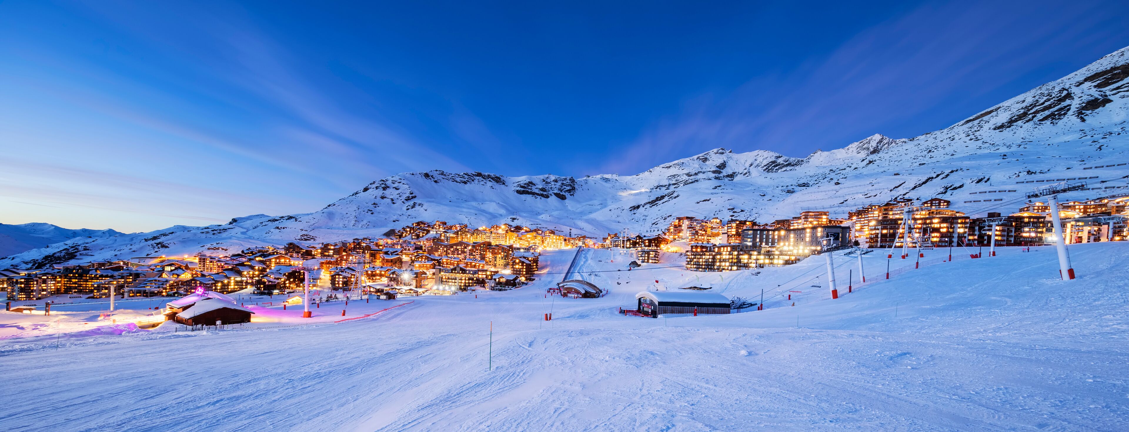 A snow-covered mountain resort at dusk with illuminated buildings and ski slopes.