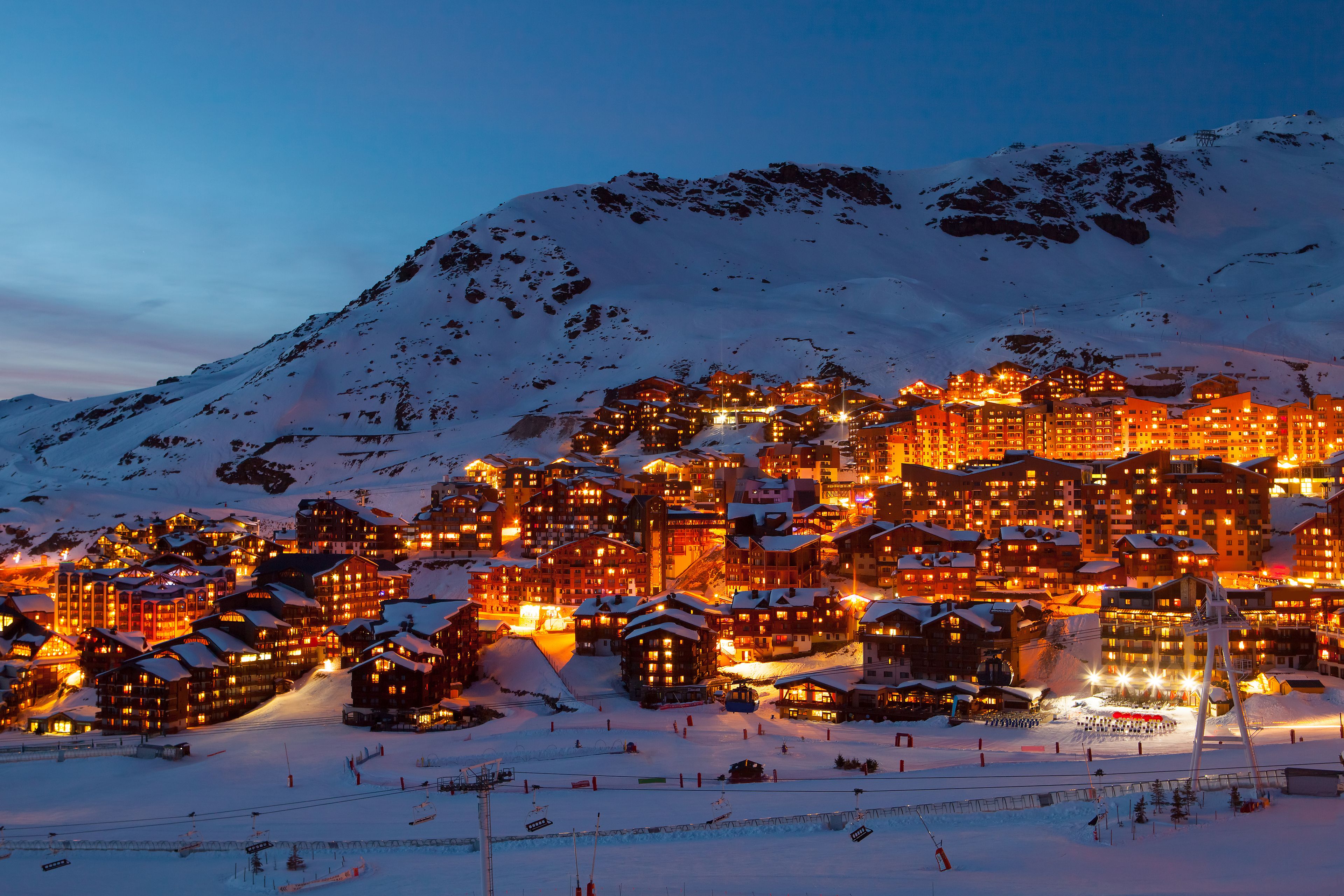 Un pueblo cubierto de nieve al anochecer, con edificios iluminados y un telesilla en primer plano.