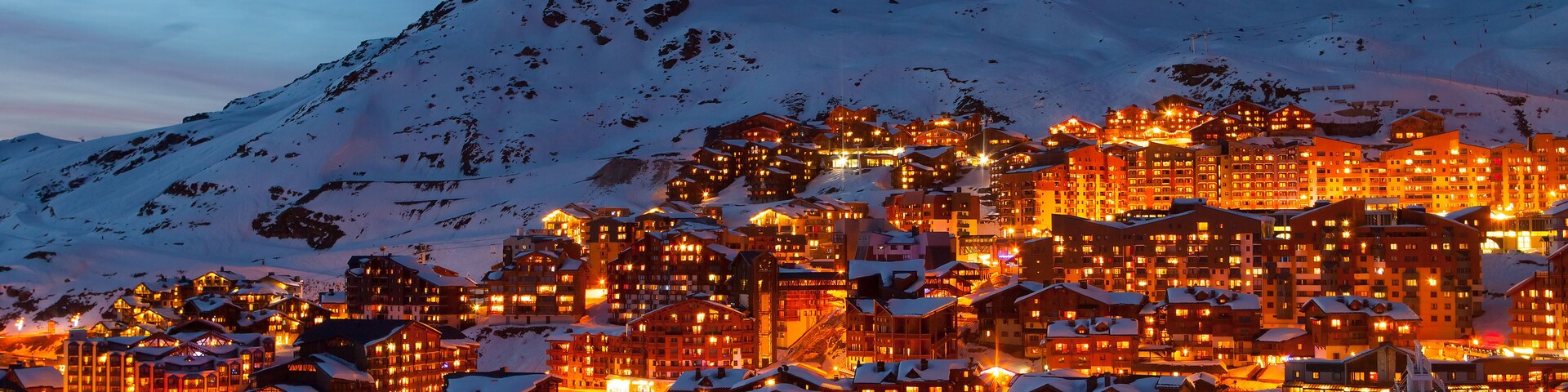 Un pueblo cubierto de nieve al anochecer, con edificios iluminados y un telesilla en primer plano.