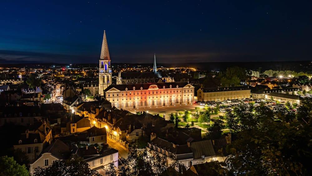 Une vue nocturne d’une ville, avec des bâtiments illuminés, un clocher d’église imposant et un grand bâtiment historique.