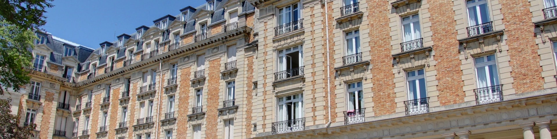 A large, historic building with multiple balconies and columns, surrounded by a well-maintained garden with sun loungers.