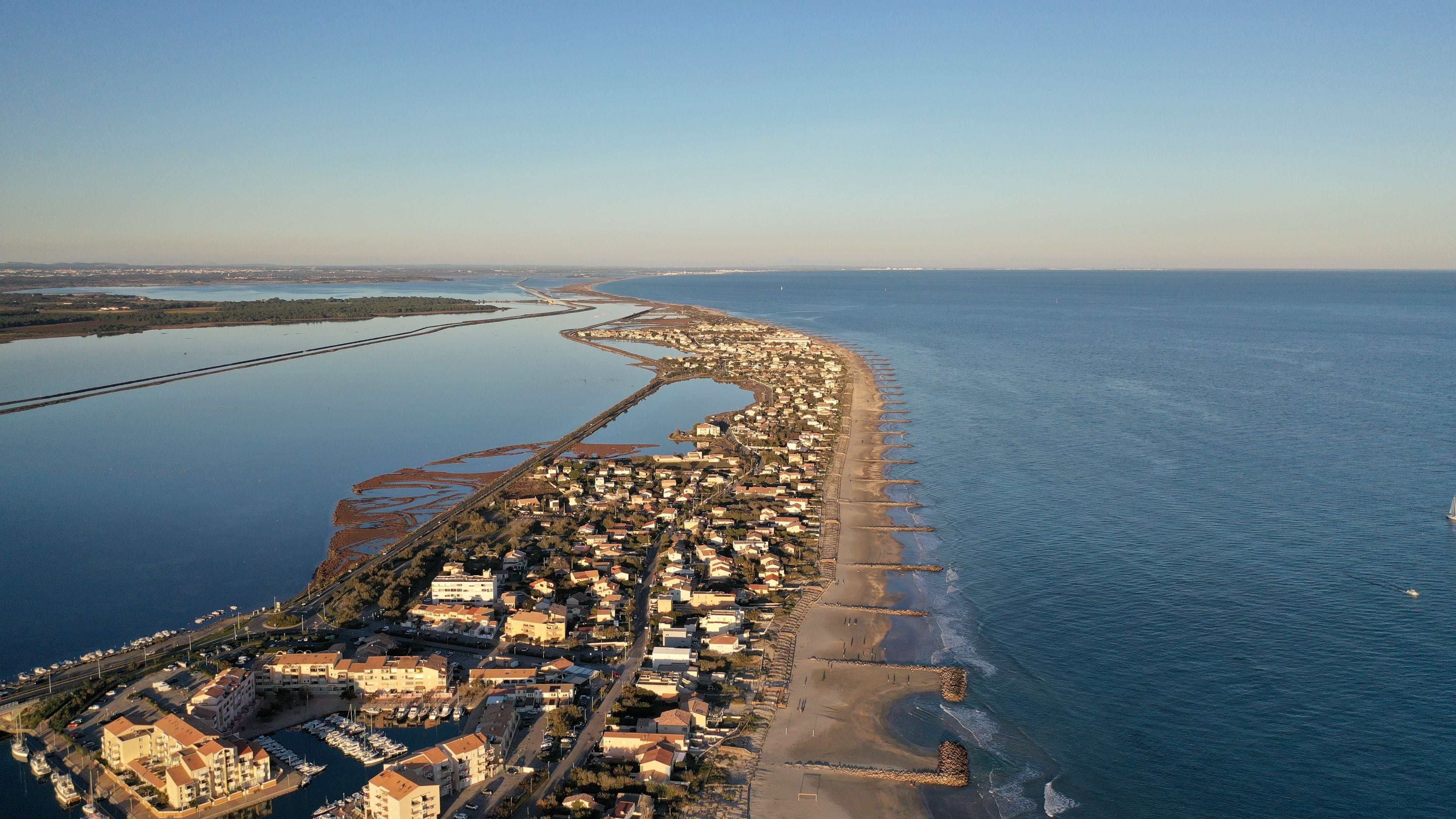 An aerial view of a coastal town with a long beachfront, a marina, and a river running parallel to the shore.