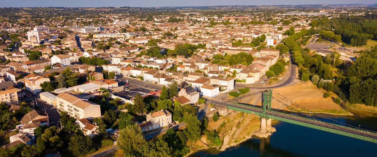 An aerial view of a town with a river, a green bridge, and surrounding greenery.