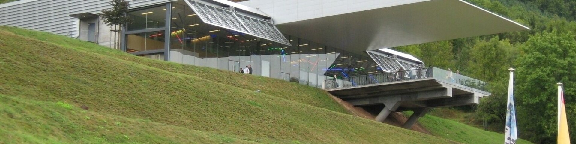 Un edificio de arquitectura moderna con techo inclinado y fachada de vidrio, ubicado en una colina cubierta de hierba con un bosque de fondo.