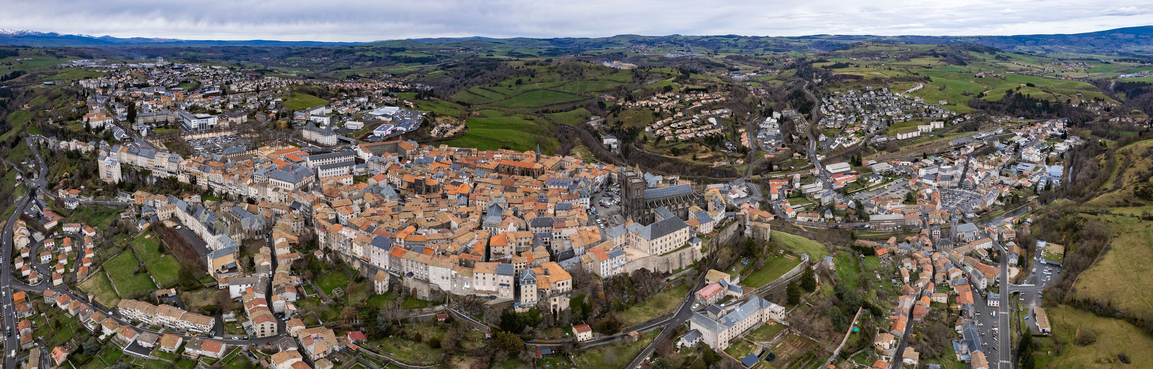 An aerial view of a historic town with a prominent castle, surrounded by rolling hills and green fields.