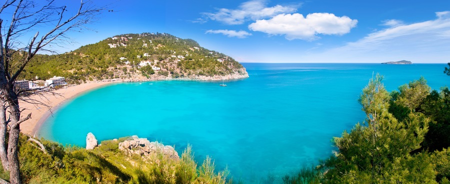 A coastal landscape with a turquoise sea, a sandy beach, and a hill with buildings.