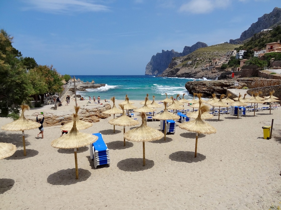 A beach with thatched umbrellas, blue lounge chairs, and rocky cliffs in the background.