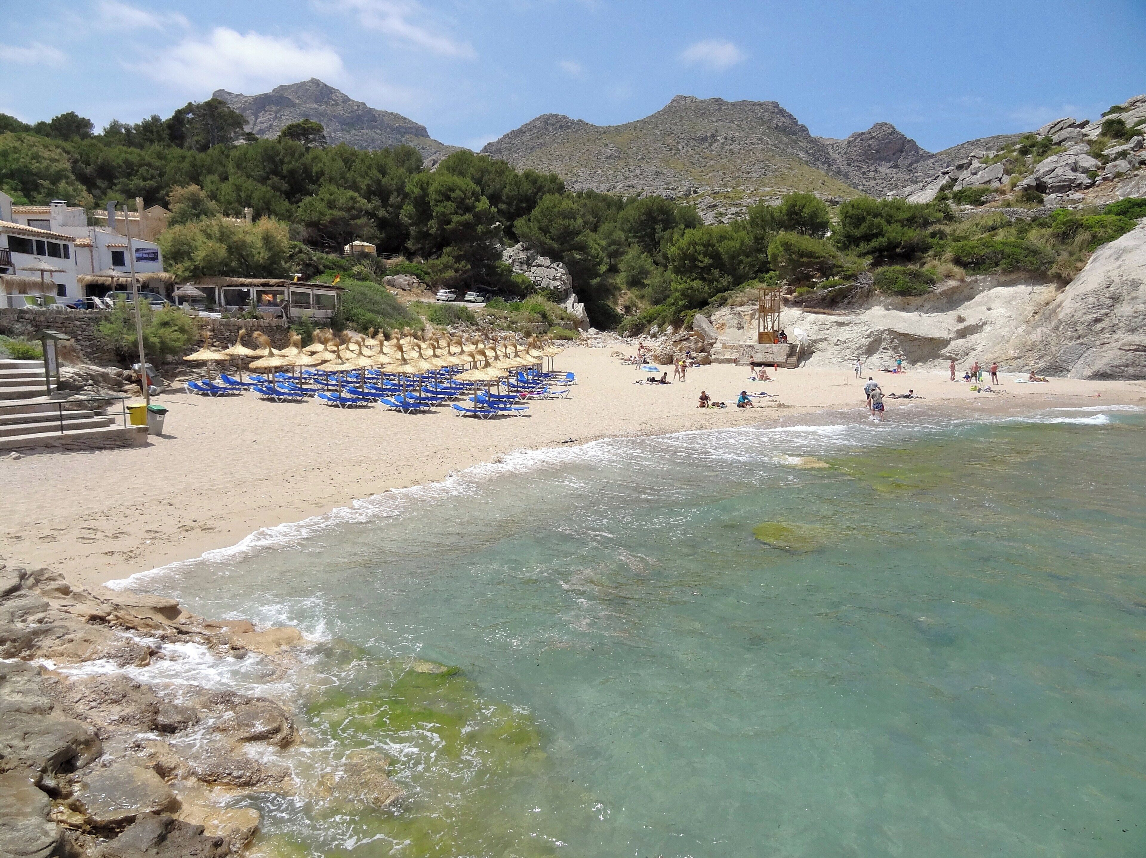 A beach with clear turquoise water, rocky shoreline, and mountains in the background.