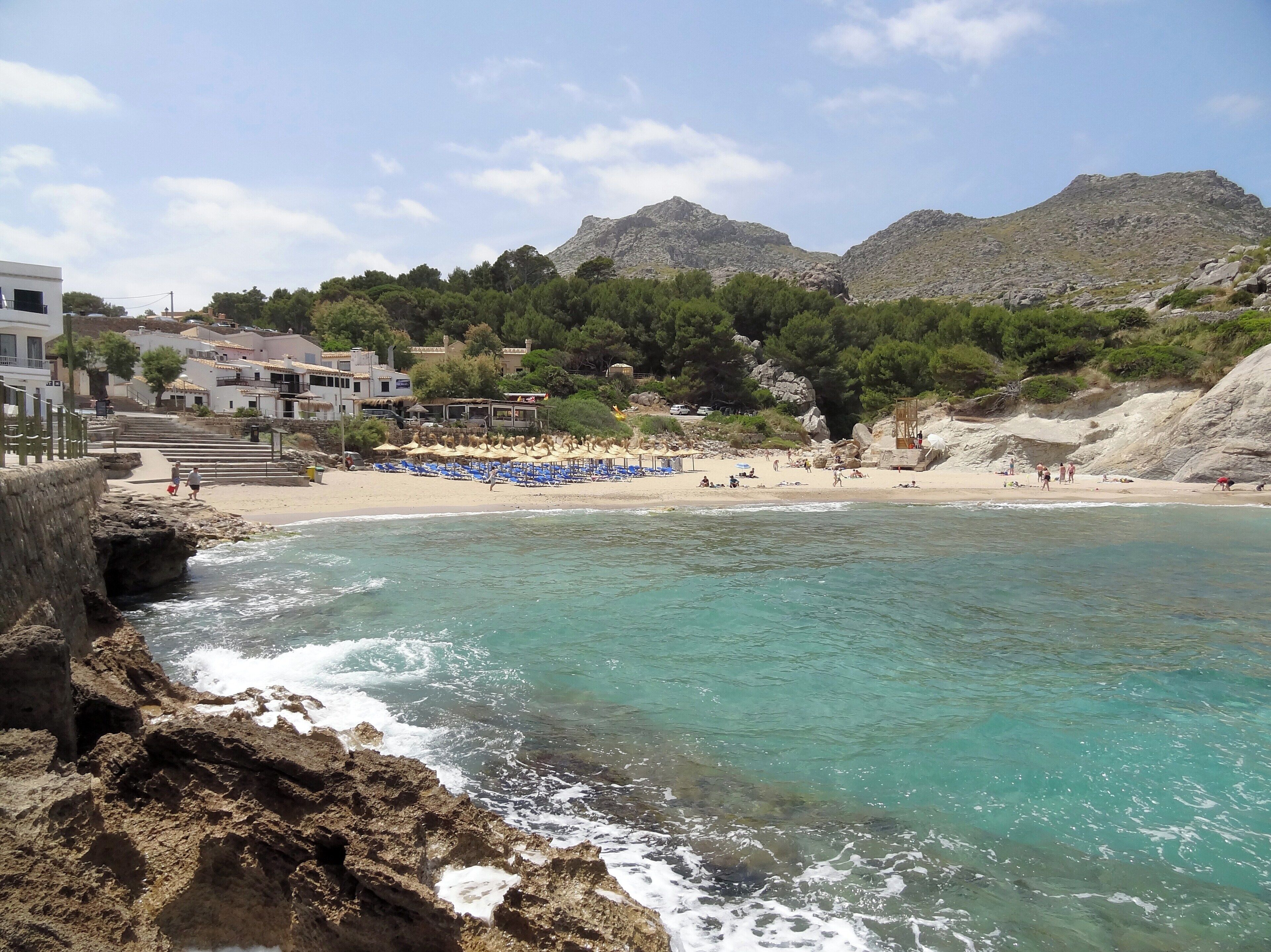 A coastal beach with clear turquoise waters, rocky cliffs, and a mountainous backdrop.