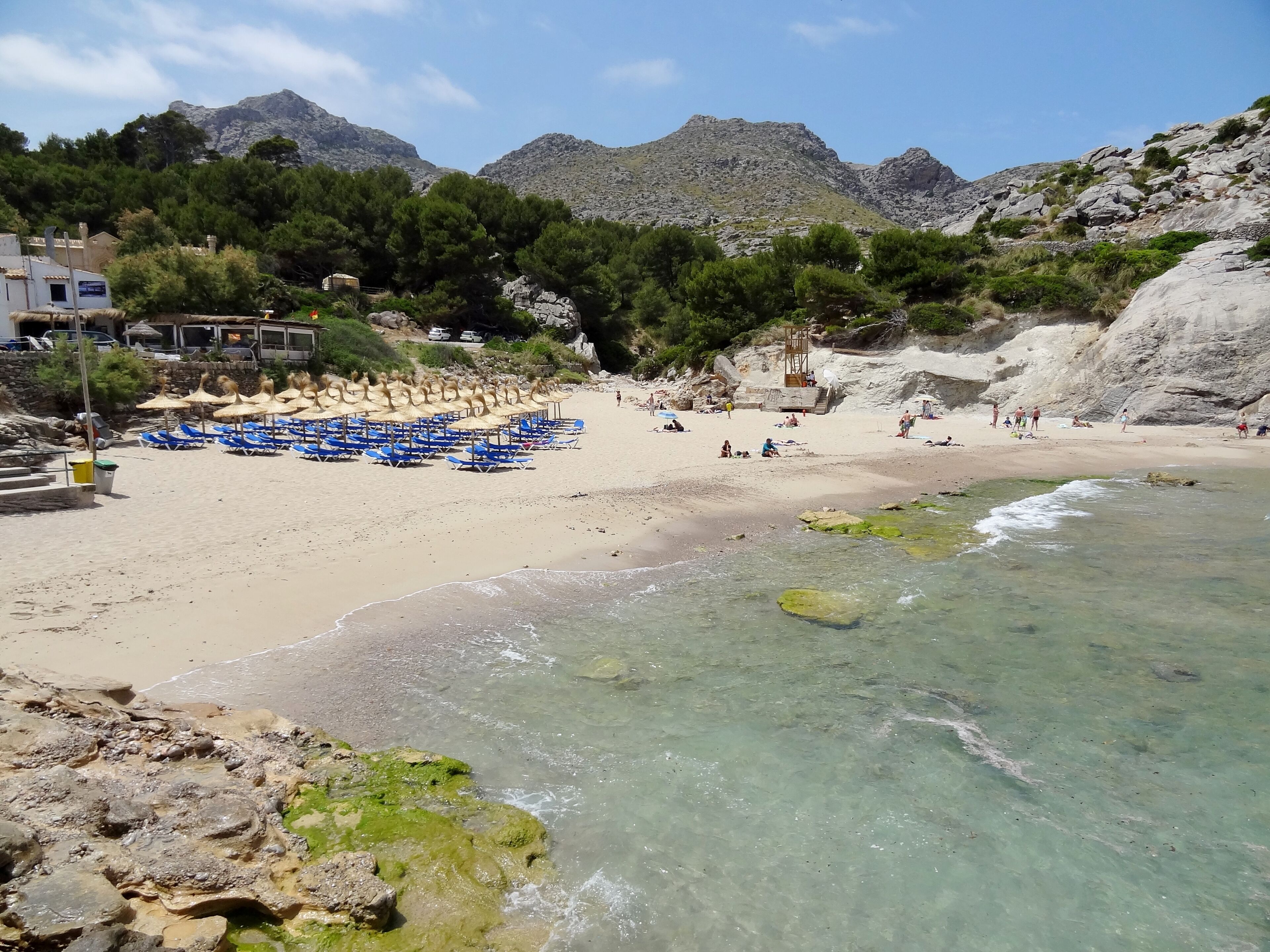 A beach with clear water, blue umbrellas, and rocky mountains in the background.