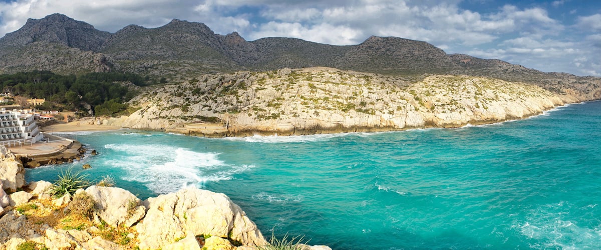 A coastal landscape with turquoise waters, rocky cliffs, and a mountainous backdrop.