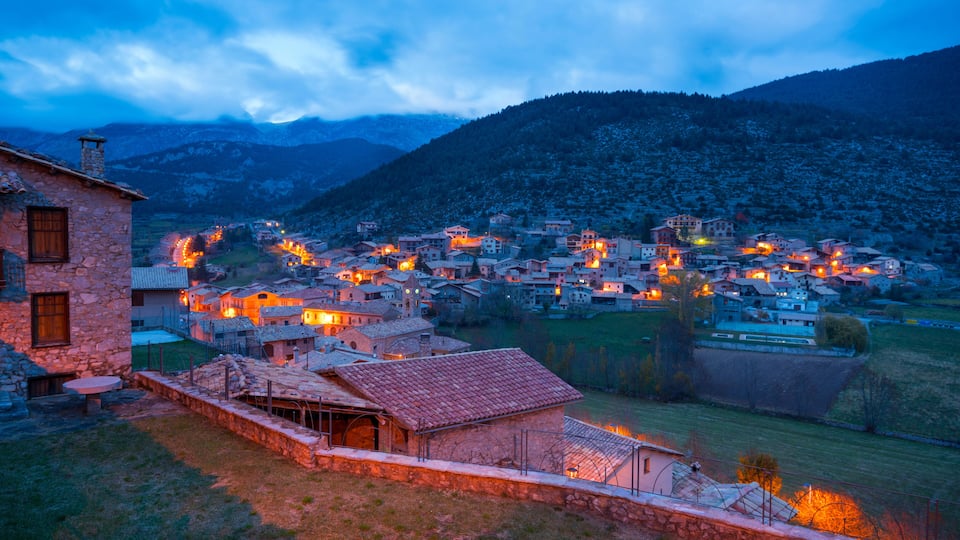 A village at dusk with illuminated buildings and a mountainous backdrop.