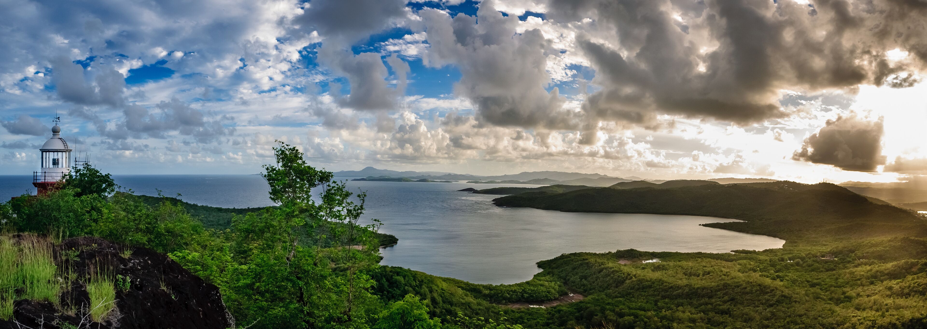 A lighthouse on a rocky outcrop overlooking a bay with lush greenery and distant mountains.