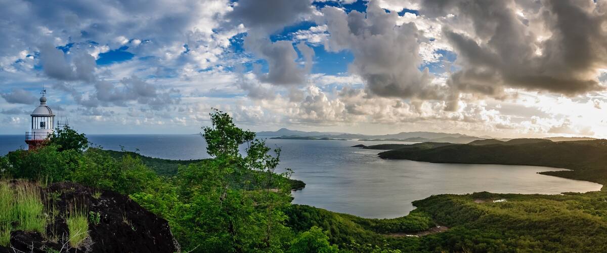 A lighthouse on a rocky outcrop overlooking a bay with lush greenery and distant mountains.