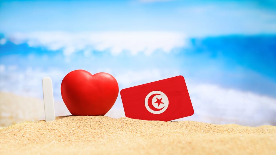 A red heart and a red card with a white crescent and star on a sandy beach with a blue sky and ocean in the background.
