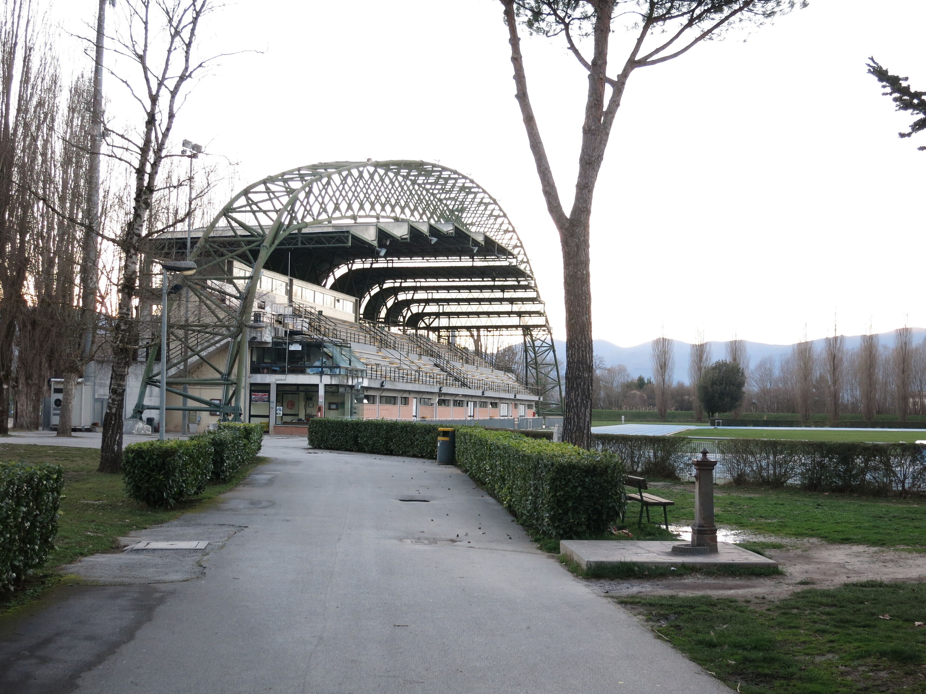 A stadium with a large, open roof structure, surrounded by trees and a pathway leading to it.