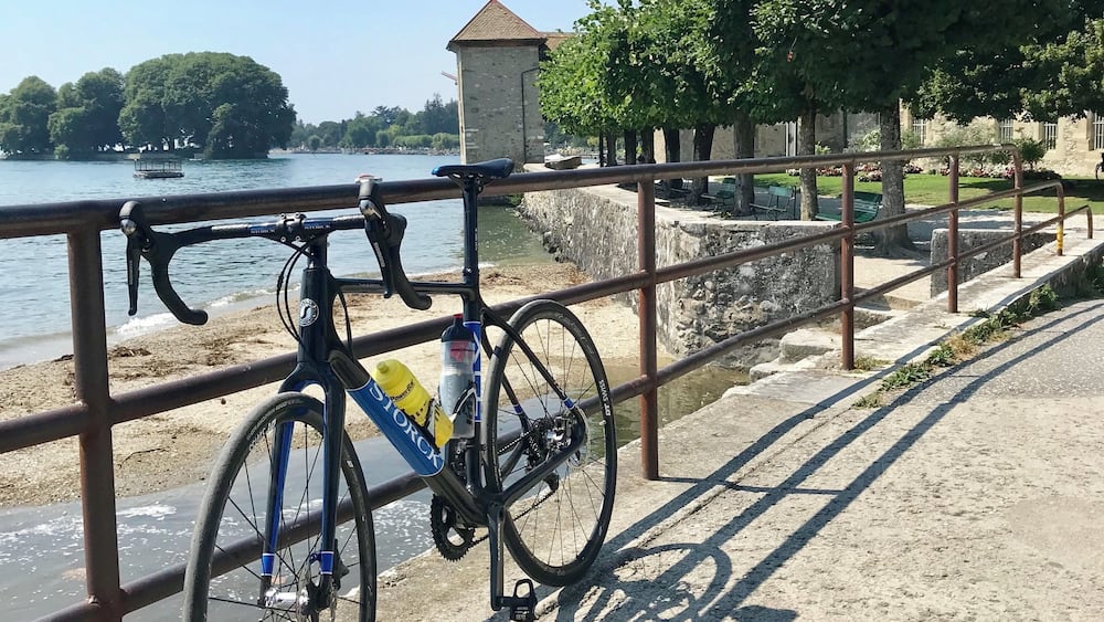 A bicycle parked by a metal railing overlooking a body of water, with a stone building and trees in the background.