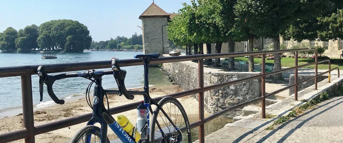 A bicycle parked by a metal railing overlooking a body of water, with a stone building and trees in the background.