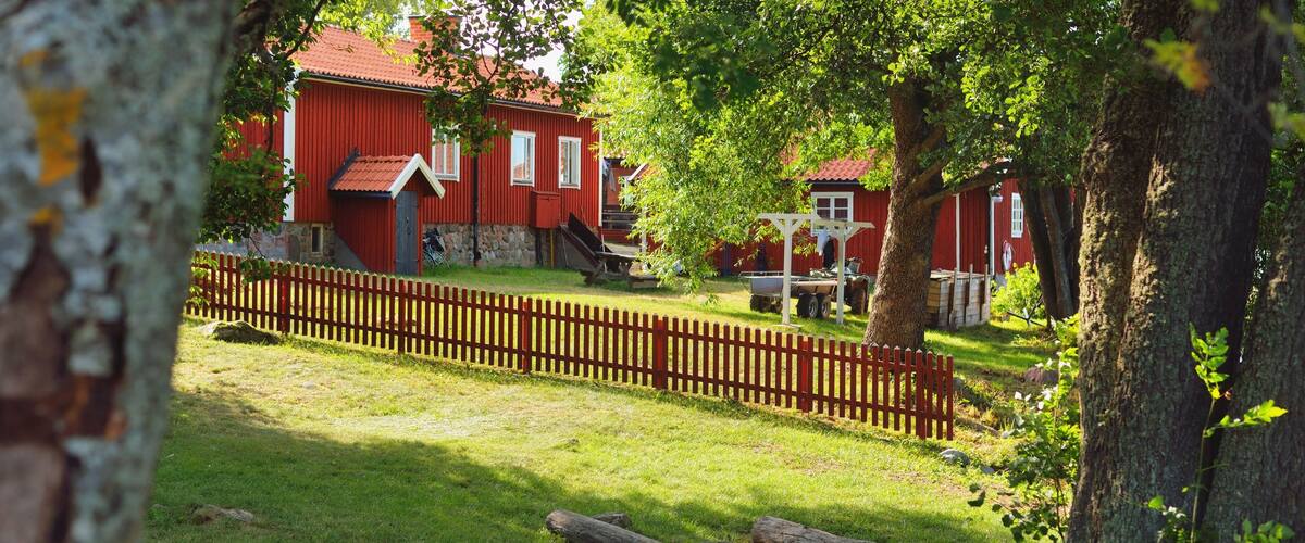 A red wooden house with a stone foundation, surrounded by a wooden fence and trees.