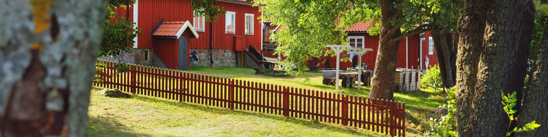 A red wooden house with a stone foundation, surrounded by a wooden fence and trees.